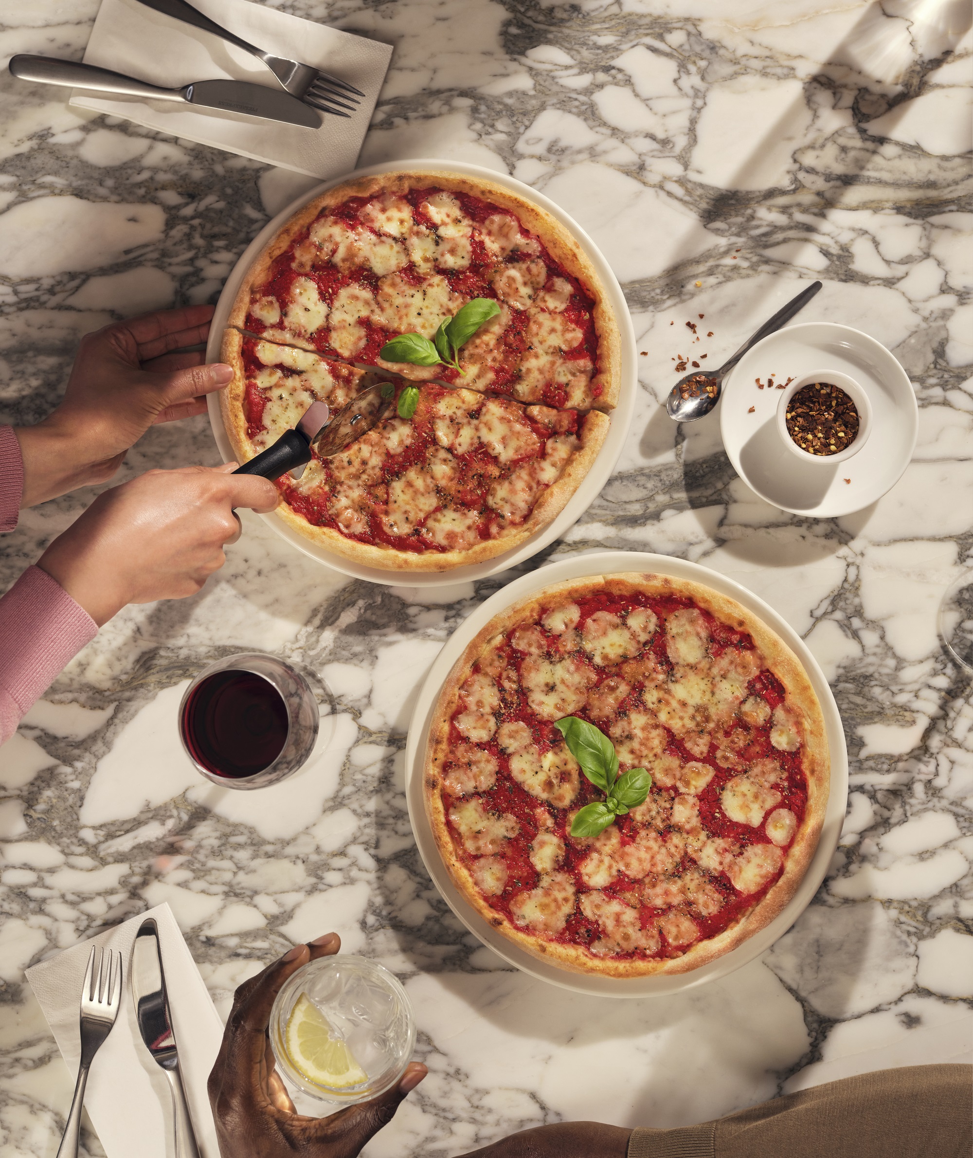 Two margarita pizzas are on a marble table with someone cutting one of them
